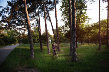 Fototapeta premium Young mother holds baby in arms in the rays of the sunset in the park between pine trees green grass. Woman kisses, hugs girl daughter. Maternal care, custody. Adoption concept. Walks in the open air