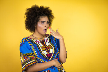 African american woman wearing african clothing over yellow background covering mouth with hand, shocked and afraid for mistake. surprised expression