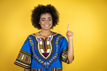 African american woman wearing african clothing over yellow background showing and pointing up with fingers number one while smiling confident and happy