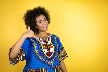 African american woman wearing african clothing over yellow background smiling doing phone gesture with hand and fingers like talking on the telephone
