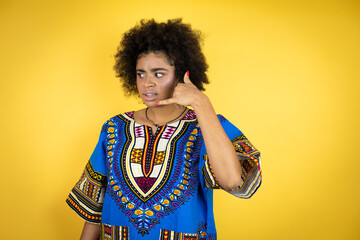 African american woman wearing african clothing over yellow background confused doing phone gesture with hand and fingers like talking on the telephone