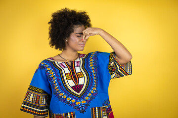 African american woman wearing african clothing over yellow background smelling something stinky and disgusting, intolerable smell, holding breath with fingers on nose