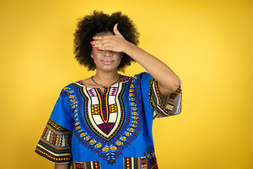 African american woman wearing african clothing over yellow background serious and covering her eyes with her hand
