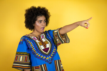 African american woman wearing african clothing over yellow background pointing with finger surprised ahead, open mouth amazed expression, something on the front