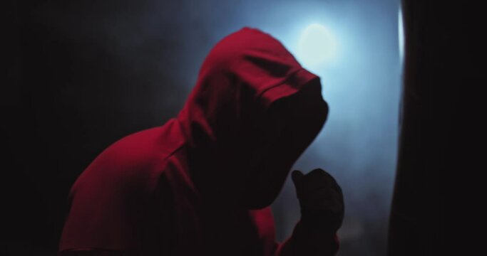 Portrait, a man fighter hits a punching bag with all his might, strong punches. Silhouette of a fighter in a hood in a dark gym, tubers of smoke. 4k, ProRes