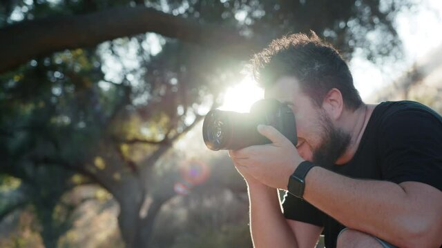 Professional photographer with sun rays on backlight excited by artistic photoshoot in nature park. Man taking pictures outdoors on summer day at sunset. Slow motion handsome man looking in pro camera