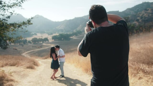 Back view of professional photographer making pictures of family couple in nature park with mountain landscape on sunny summer day. Lovers kissing in front of camera. Photographer shooting models