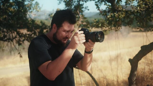 Slow motion handsome man using pro camera for photoshoot. Front view of professional photographer face looking on the camera screen and taking pictures in the nature park on sunny summer day, 4K