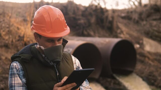 portrait man with a digital tablet is exploring environmental pollution radiation waste. a man in a respirator mask and an orange helmet stands next to the waste pipes. nature conservation protection