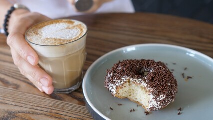 Hand taking a glass of plant based cappuccino from the table. Bitten doughnut on a plate.