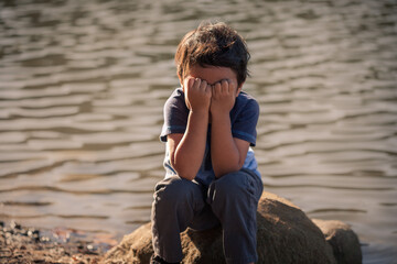 A young boy by the edge of a lake covering his face, body language expressing warning signs of emotional child abuse and isolation.