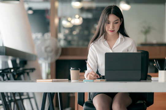 Portrait of attractive woman siiting at office desk, writing on notebook and looking at laptop screen.