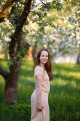 Fototapeta na wymiar Young woman in beige dress in a blooming apple garden. Spring story. Brown-haired girl with long hairs. Woman plays with her hairs