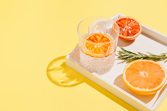 Half of grapefruit and bloody orange, rosemary on a white wooden tray and glass with water on bright yellow background. Summer refreshment concept. Sunlit flat lay. Minimal style. Top view.