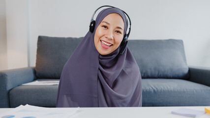 Asia muslim lady wear headphone using computer laptop talk to colleagues about plan in video call meeting while remotely work from home at living room. Social distancing, quarantine for corona virus.