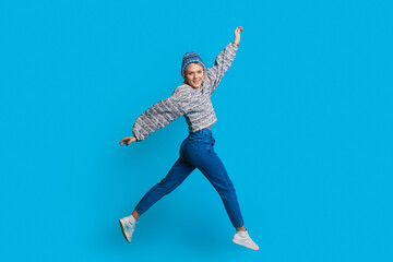 Fototapeta na wymiar Blonde woman with warm hat is jumping on a blue studio wall smiling at camera