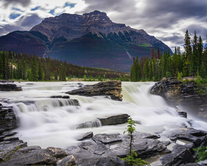 Fototapeta na wymiar The roaring waters of Athabasca Falls comes from a melting mountain glacier shown in the background. Travel along the Icefields Parkway in Jasper Park, Canada during the summer