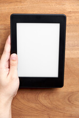 Left hand holding an electronic reader with blank screen on a wooden desk