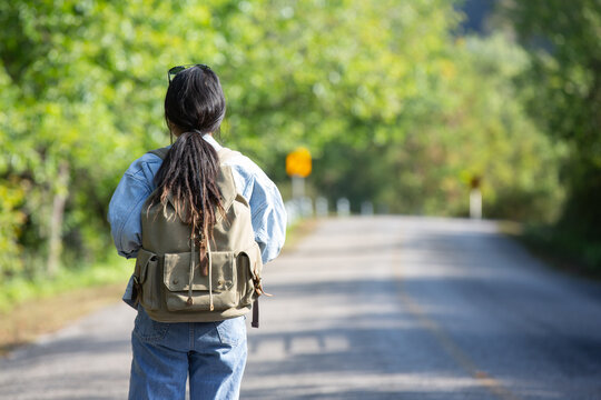 woman and backpack viewed from back and long asphalt road staight in the middle of a high trees nature forest