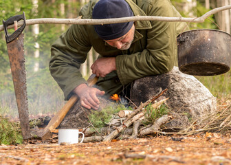 man in the forest trying to light a campfire, a tree branch with a pot over the campfire, blurred forest background, bonfire and smoke, autumn time