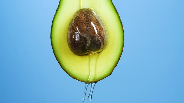 Slow motion close-up of fresh avocado fruit and oil flowing over blue background