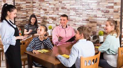 Adults with kids are giving order to cheerful waitress in comfy cafe.