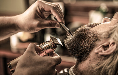 Man visiting hairstylist in barbershop. Barber works with a beard clipper. Hipster client getting haircut - obrazy, fototapety, plakaty