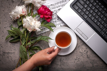 Coffe, peonies and laptop on grey vintage background. Women hand holds cup of coffee.