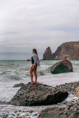 A beautiful girl in a white shirt and black swimsuit stands on a rock, big waves with white foam. A cloudy stormy day at sea, with clouds and big waves hitting the rocks.