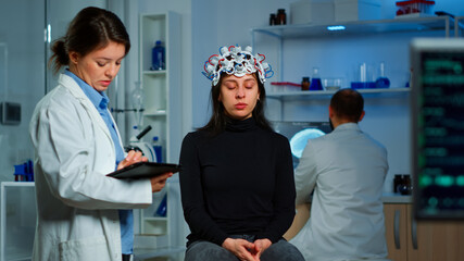 Fototapeta na wymiar Stressed patient sitting on neurological chair with eeg headset, researcher examining health status writing on tablet. Medical scientist looking through microscope in background developing treatment