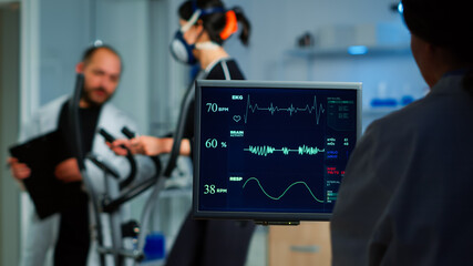 Medical researcher examining EKG image showing on monitor while patinet with mask running on cross trainer testing heart rate using electrodes.Doctor monitoring physical endurance in science sport lab © DC Studio