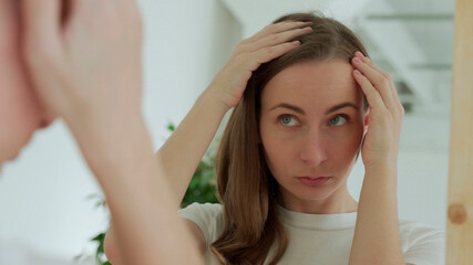 Fototapeta premium Sad upset woman standing by mirror looking at matted fragile strand in her hand thinking about natural medicinal shampoo restoring hair structure