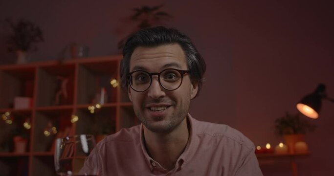 Crop view of handsome talkative man in glasses holding wineglass and talking. Cheerful male person having online communication and looking to camera while sitting at home