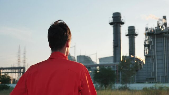 Rear View of engineer wearing safety uniform and taking off white safety helmet Looking at Industrial Factory, power plant during sunset. Slow motion