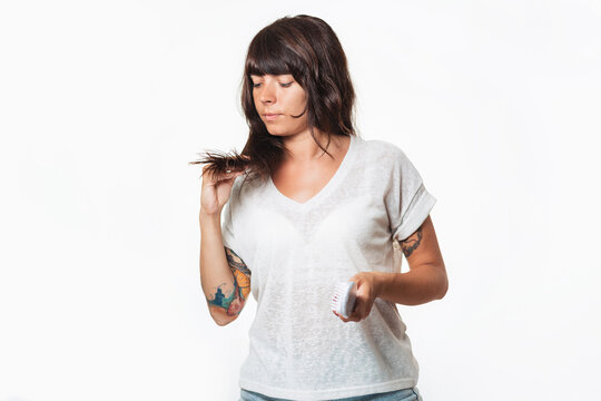 A woman with a tattoo on her arm holds a comb, and examines the tips of her hair. Copy space. White background. Hair care concept