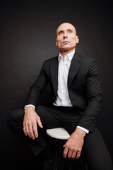 Portrait of confident elegant businessman in black suit sitting on stool and looking away