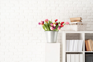 Bucket of tulip flowers next to the bookshelf over white brick wall background