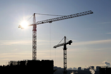 Silhouettes of construction cranes and unfinished residential building against the sky and shining sun. Housing construction, apartment block in city