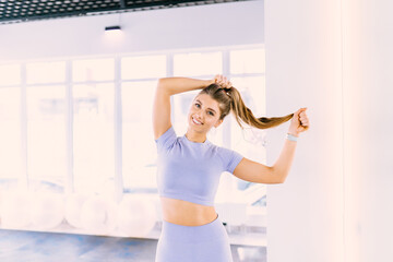 Fototapeta na wymiar Portrait of trainer fitness woman in sports clothing looking confident. Young female wearing light lilac sportswear. Female model with perfect tanned body. Smiling female correct hair near mirror.