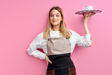 nice woman waitress in apron, offering cup of delicious tasty coffee on tray, stand smiling, friendly staff of restaurant. isolated over pink studio background - obrazy, fototapety, plakaty