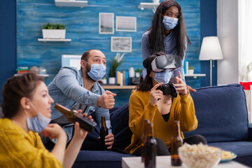 Fototapeta na wymiar Man with face mask cheering up for friends playing video games with vr headset in home living room keeping social distancing in time of global pandemic with covid. Multi ethnic people having fun.