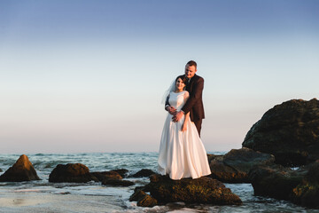 Fototapeta na wymiar the groom and the bride stand on a stone in the sea at sunset. beautiful wedding couple at sunset of the day is on the beach. beautiful beach with gentle waves during sunset with newlyweds in the back