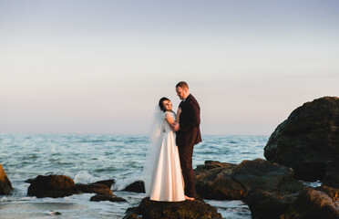 the groom and the bride stand on a stone in the sea at sunset. beautiful wedding couple at sunset of the day is on the beach. beautiful beach with gentle waves during sunset with newlyweds in the back