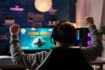 Excited player with headphones making winner gesture while playing games on computer at gaming home studio. Online streaming cyber performing gaming tournament using technology network wireless