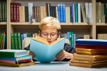 clever boy reading book in library, keen on education, preparing for school, wearing eyeglasses
