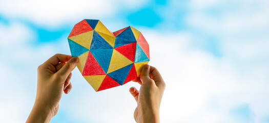 Child hands holding colorful heart over blue sky background. World autism awareness day concept