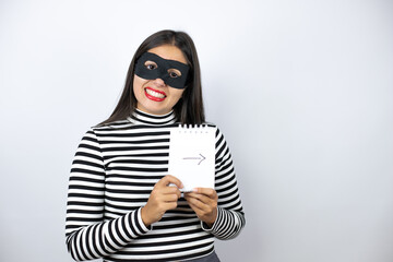 Young beautiful brunette burglar woman wearing mask holding a notebook with a arrow drawn on it