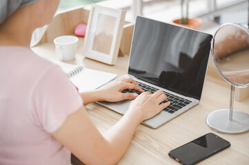 Naklejka premium Young Asian woman wearing a hijab uses smartphone and laptop at her desk. Attaching a pink ribbon represents recovery from a breast cancer patient. Breast cancer concept, cancer prevention concept.