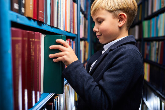 Child Boy Is Searching And Choosing Book In Bookshop, Stand Near Shelves, Going To Learn And Study. Side View