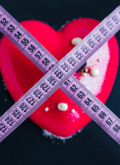 No sweets. High-calorie cake in the shape of a red heart on a black background crossed out with a measuring tape